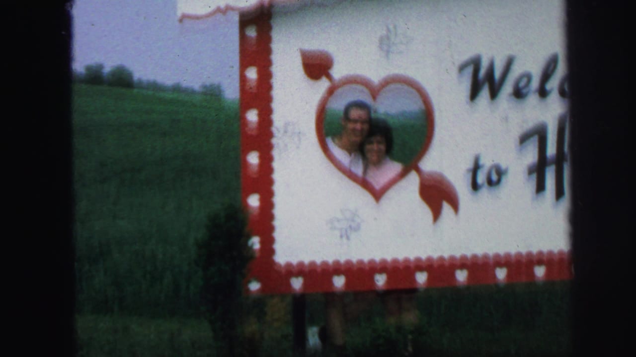 Adult couple smiling through a red heart cutout on a white welcome sign beside a grassy hill.