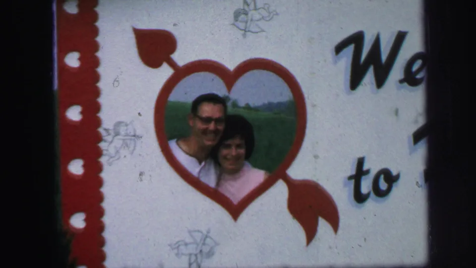 A closer 8mm frame shows the couple smiling inside the red heart cutout, with partial welcome lettering and faint cherub sketches on the sign.