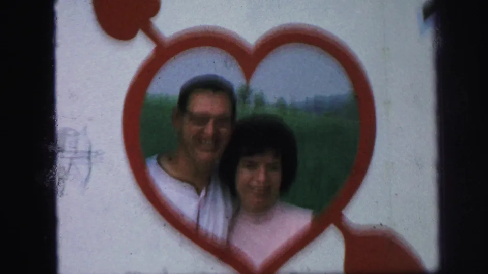 A tight 1971 home-movie frame centers the adult couple behind the red heart shape, with the grassy background visible through the opening.