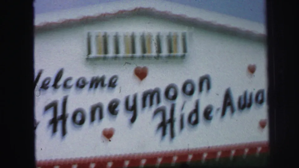 The final frame centers on the painted Welcome to Honeymoon Hide-Away sign, with red hearts and a filmstrip border on the white wall.
