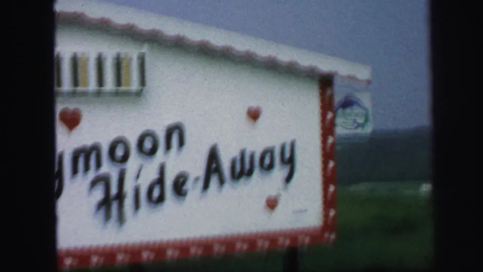 1971 Lynbrook home-movie frame showing a white and red Honeymoon Hide-Away sign, partly cropped, with hearts and dark film edges.