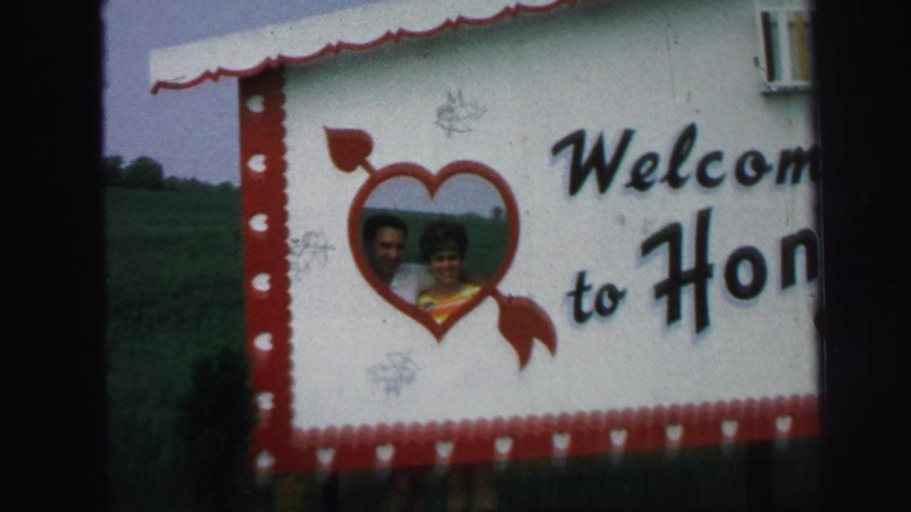 1971 Lynbrook frame of a Welcome to Honeymoon Hide-Away sign with a red heart graphic and a smiling adult couple pictured inside it.