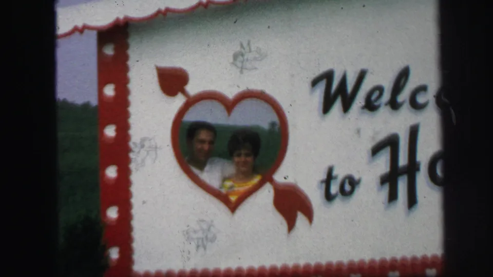 1971 Lynbrook home-movie still showing the Honeymoon Hide-Away welcome sign, red border, heart graphic, and an adult couple in the center.