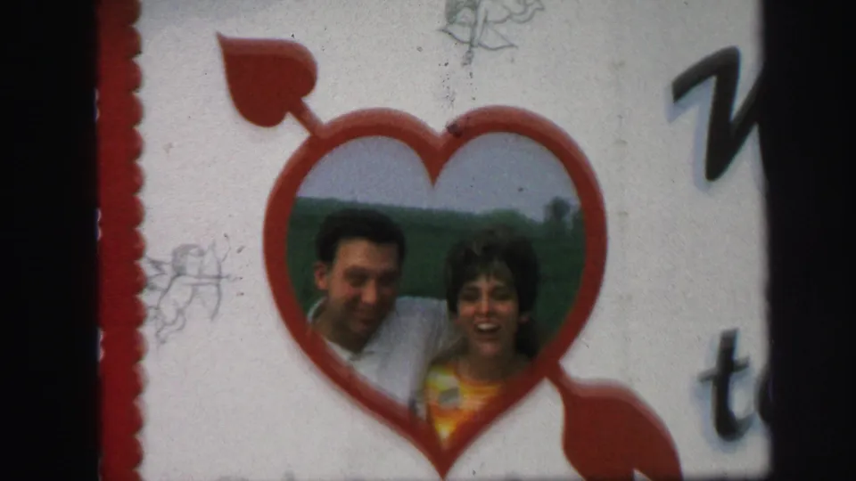 Tight 1971 Lynbrook 8mm frame of the Honeymoon Hide-Away sign's red heart graphic, with a smiling adult man and woman visible inside.