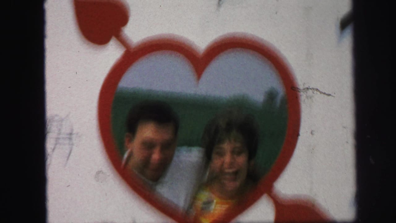 Couple smiling inside a red heart-shaped frame with green field behind them.