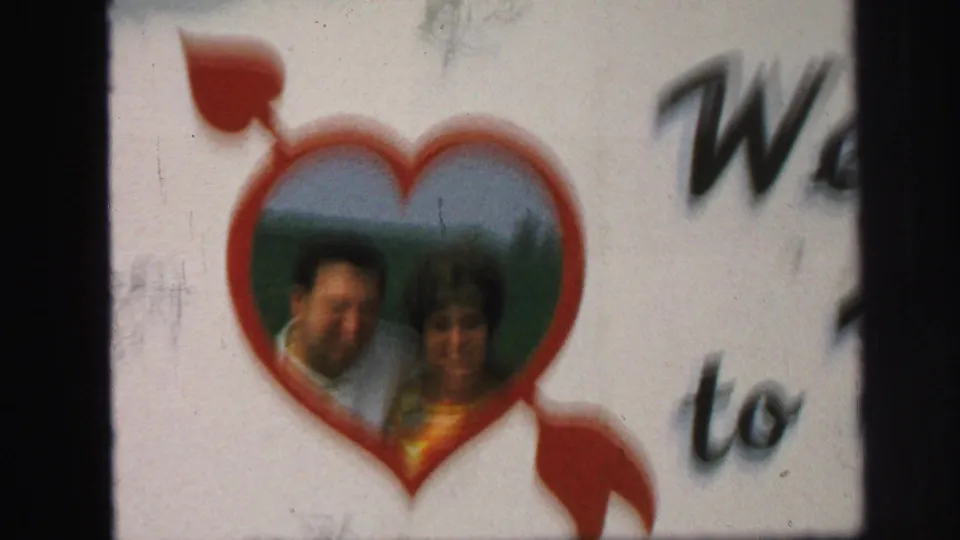 The handheld camera widens to show the couple inside the heart cutout and partial welcome text on the sign.