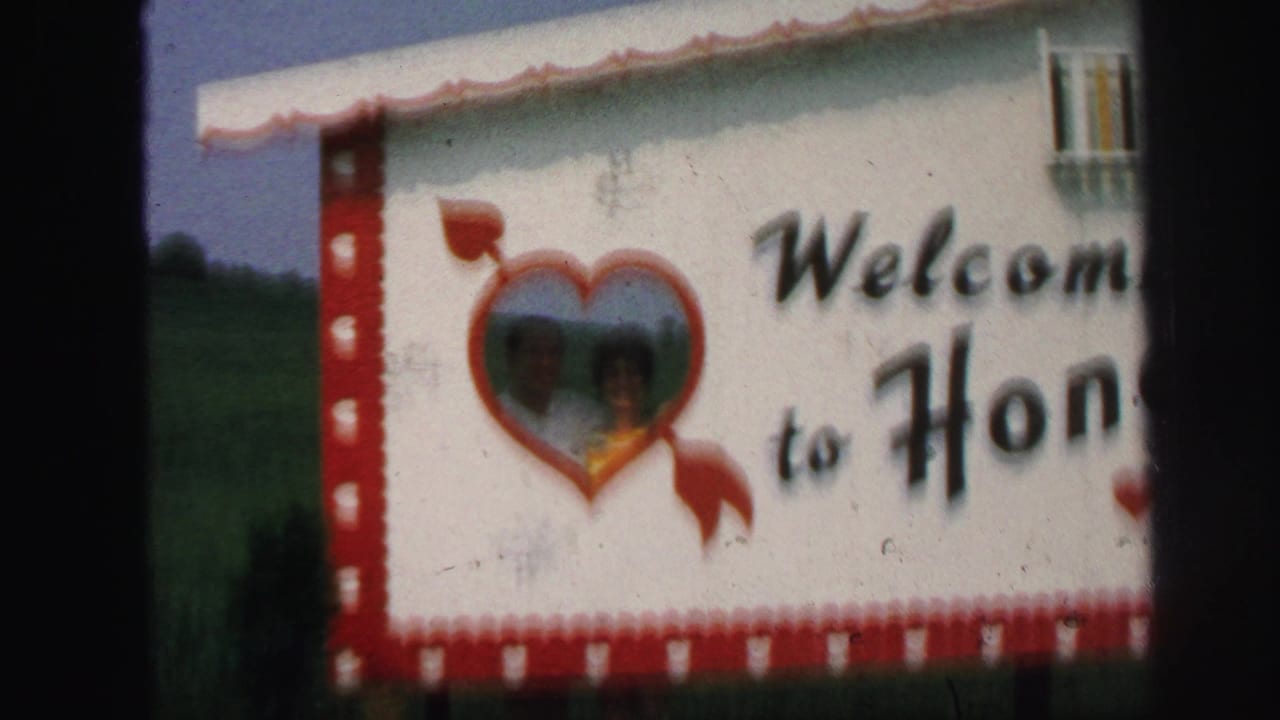 White welcome sign with red border and heart-shaped opening beside a grassy field.