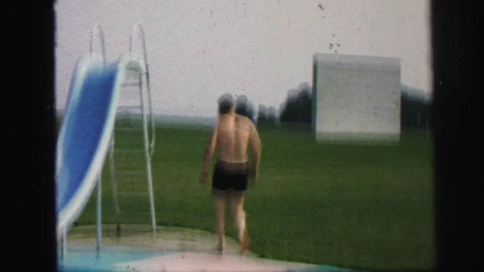 1971 Lynbrook home-movie frame showing a man in dark swim trunks walking barefoot across a lawn toward a blue outdoor pool slide.
