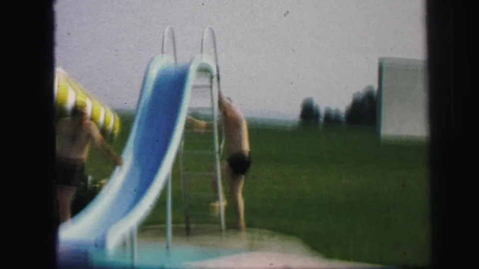 1971 Lynbrook frame showing men near a blue pool slide and ladder, with open grass and a pale rectangular structure in the background.