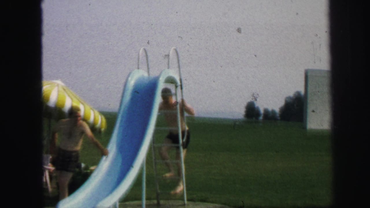 In Lynbrook, New York, 1971, a child climbs the ladder of a blue pool slide while a striped umbrella stands nearby.