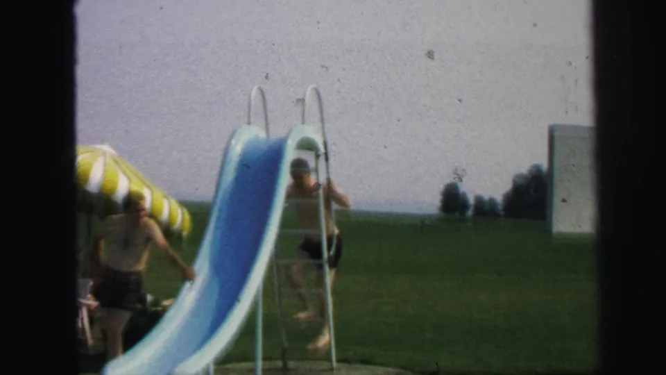In Lynbrook, New York, 1971, a child climbs the ladder of a blue pool slide while a striped umbrella stands nearby.