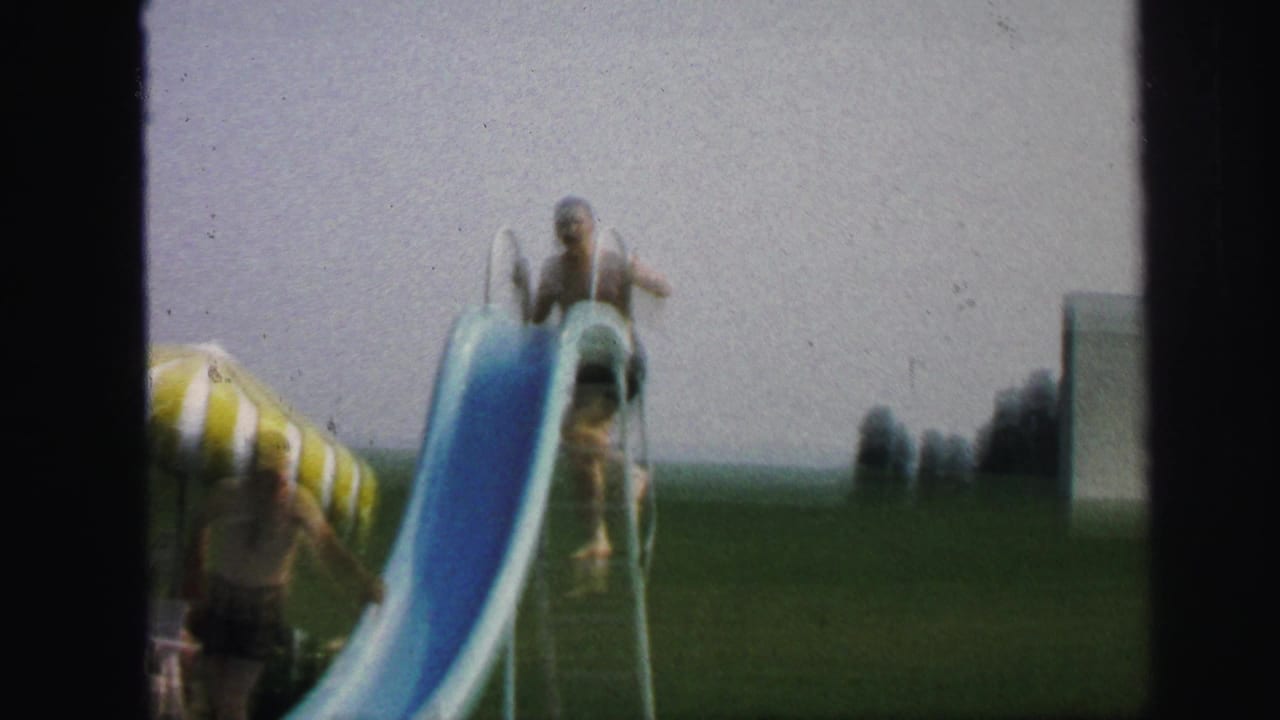 In the 1971 Lynbrook backyard pool scene, a child reaches the top of a blue slide under a pale sky.