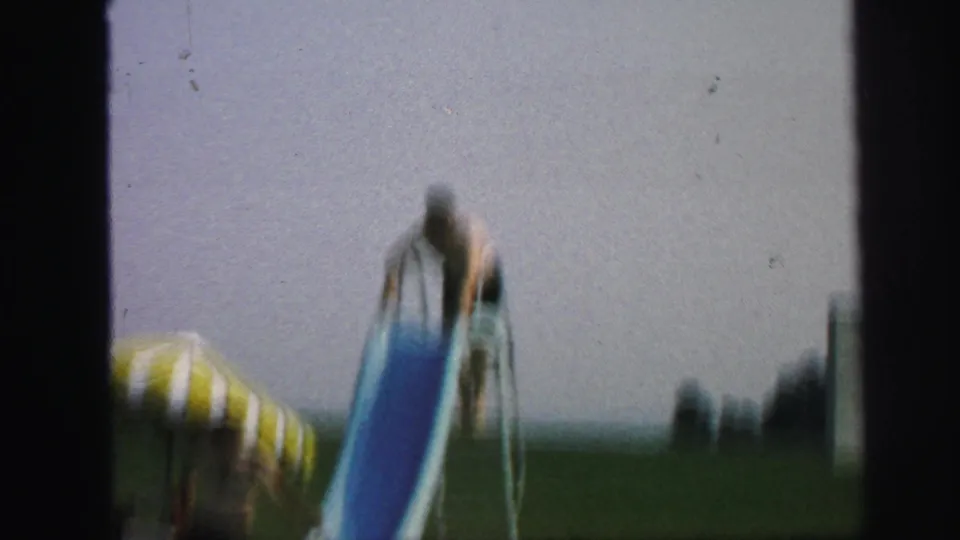A child starts down the blue pool slide in a soft, grainy 1971 home-movie frame from Lynbrook, New York.