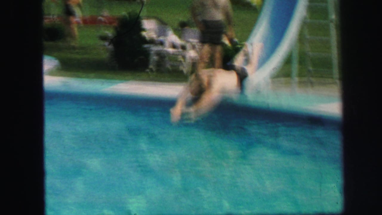 In Lynbrook, New York, 1971, a swimmer in dark trunks descends a pale backyard pool slide beside lawn furniture.
