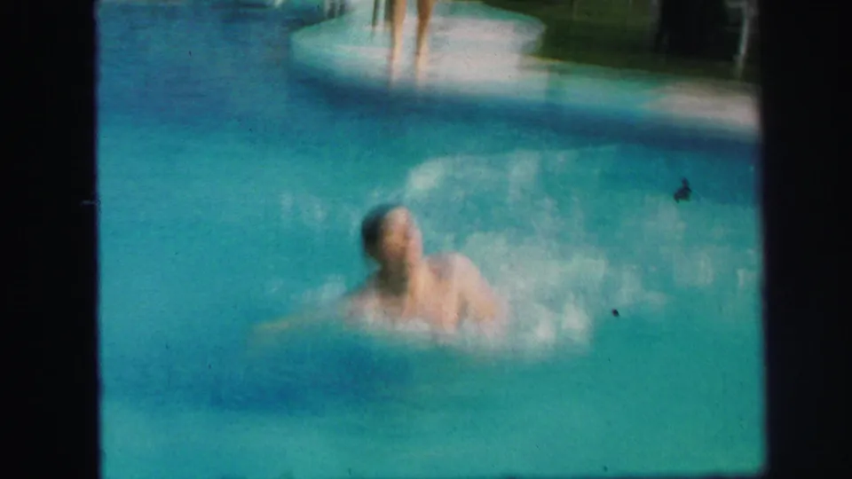 A swimmer surfaces in a backyard pool with a large splash near the pale pool edge in the 1971 Lynbrook home movie footage.