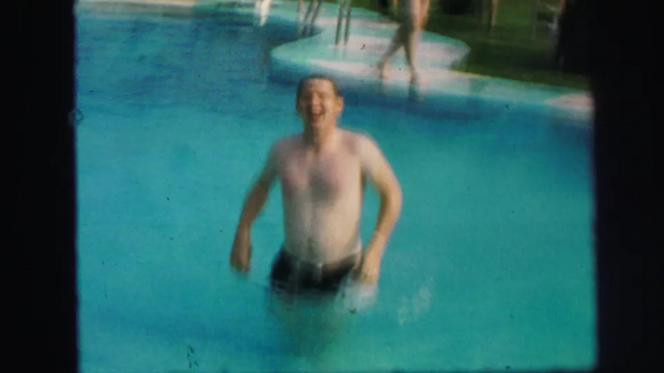 A smiling swimmer stands in turquoise pool water with a striped umbrella and pool ladder visible behind him in Lynbrook, 1971.