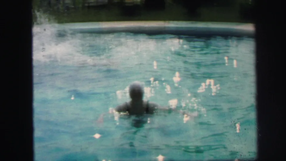 A swimmer seen from behind moves through the pool water, with bright reflections and the curved pool edge visible in the frame.