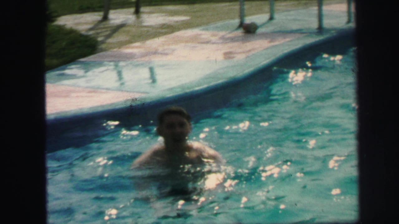 1971 Lynbrook home-movie frame showing a person in a backyard swimming pool near the deck, with soft 8mm color and grain.