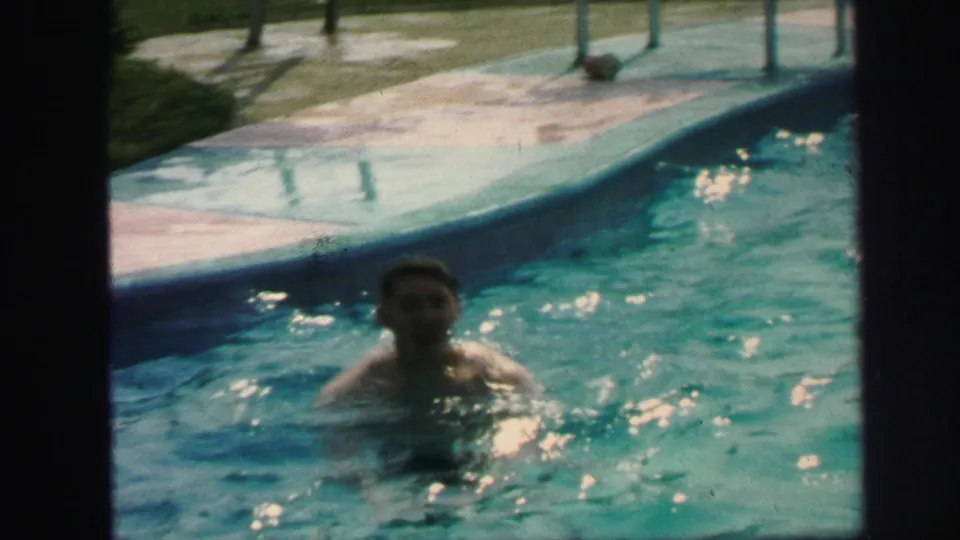 1971 Lynbrook home-movie frame showing a person in a backyard swimming pool near the deck, with soft 8mm color and grain.