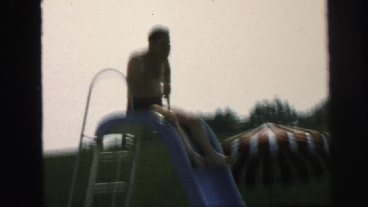 Boy in swim trunks sits on a blue pool slide in the Lynbrook backyard pool area, with a striped sunshade in the background.
