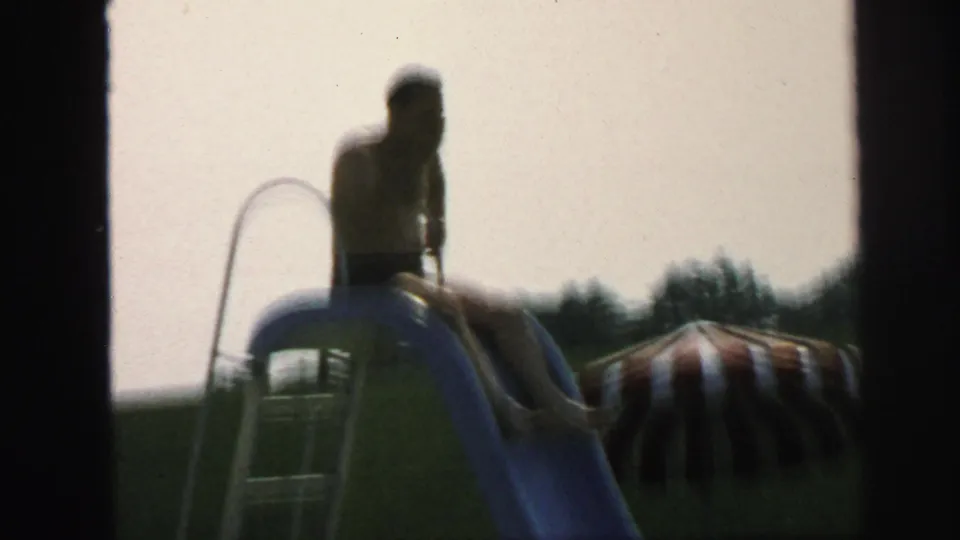 Boy in swim trunks sits on a blue pool slide in the Lynbrook backyard pool area, with a striped sunshade in the background.