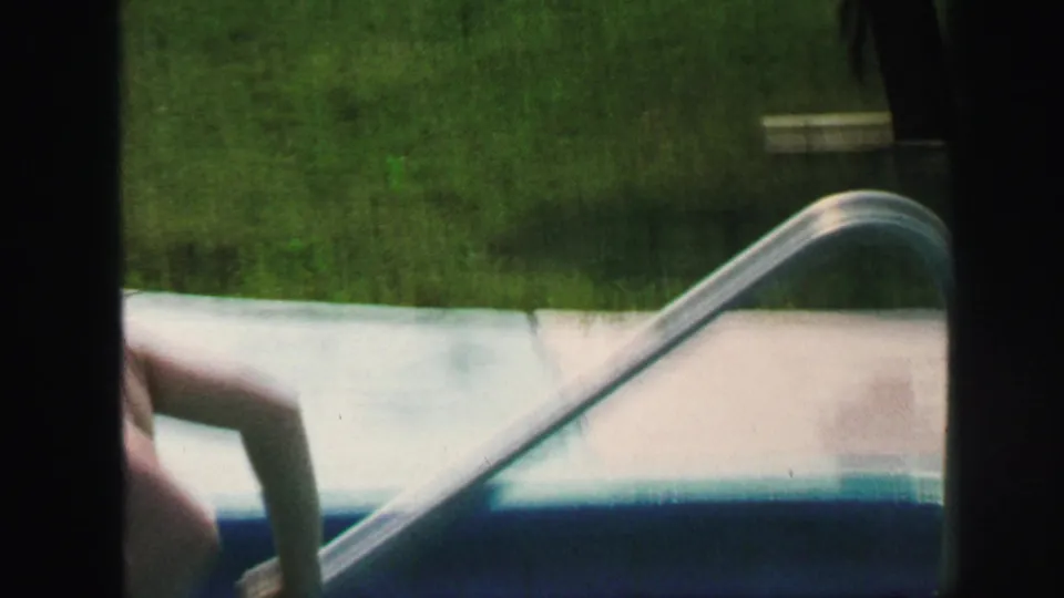 1971 Lynbrook home-movie frame showing a metal pool ladder, pale pool edge, and surrounding backyard grass.