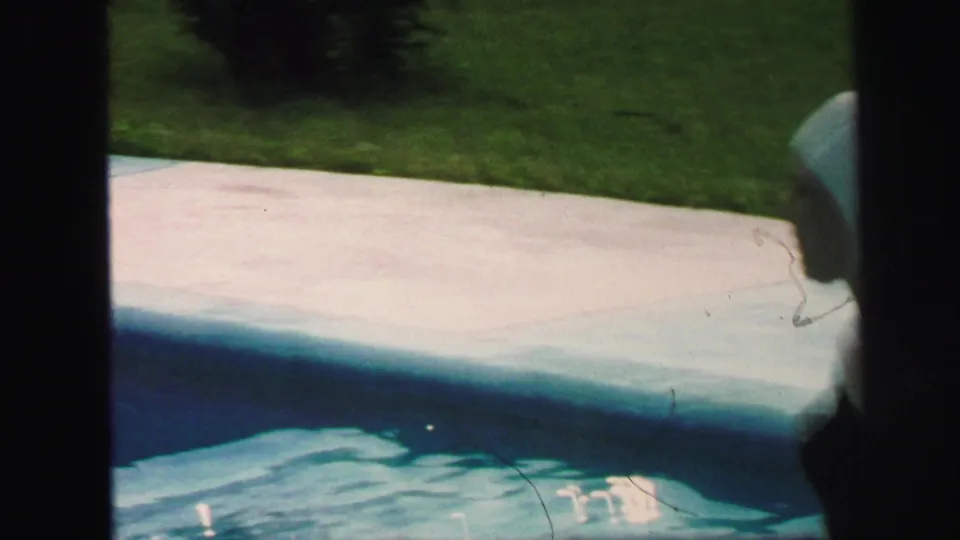1971 Lynbrook poolside frame showing the pool edge, blue water, concrete or pale deck surface, and lawn beyond.