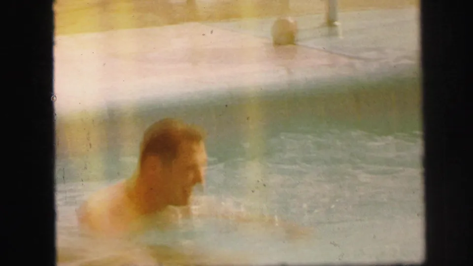 1971 Lynbrook home-movie frame with a soft, partially visible swimmer in a backyard pool near the pale concrete edge.