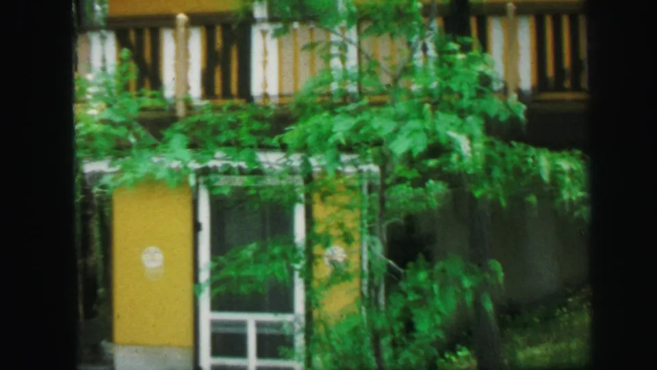 1971 Lynbrook exterior view of a yellow door and white-trimmed building partly hidden by green foliage.