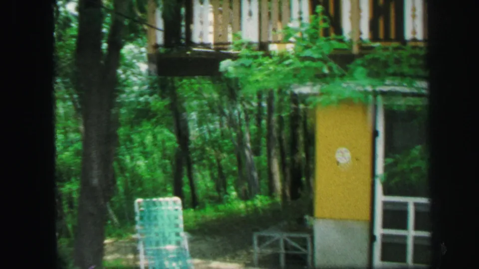 Wooded Lynbrook exterior with a yellow door, white trim, trees, and a folding lawn chair in the yard.