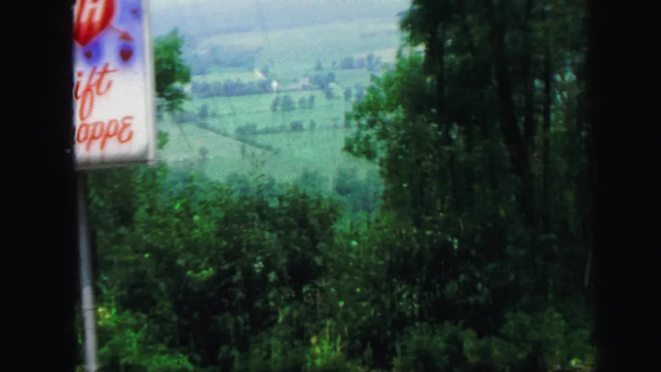 1971 8mm frame with a cropped roadside sign, wooded foreground, and rolling green fields in the distance.