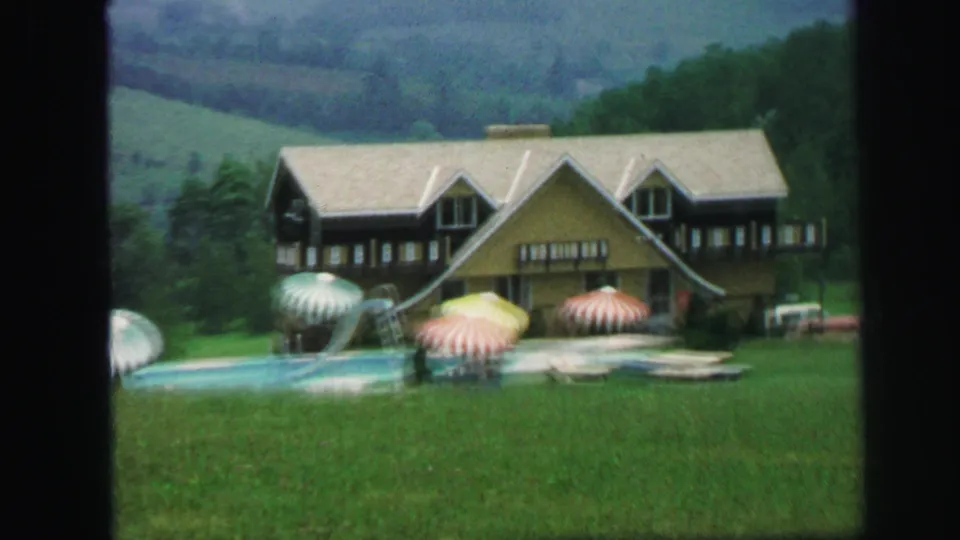 1971 8mm frame showing a large yellow and brown house, pool area, colorful umbrellas, lawn, and wooded hills.