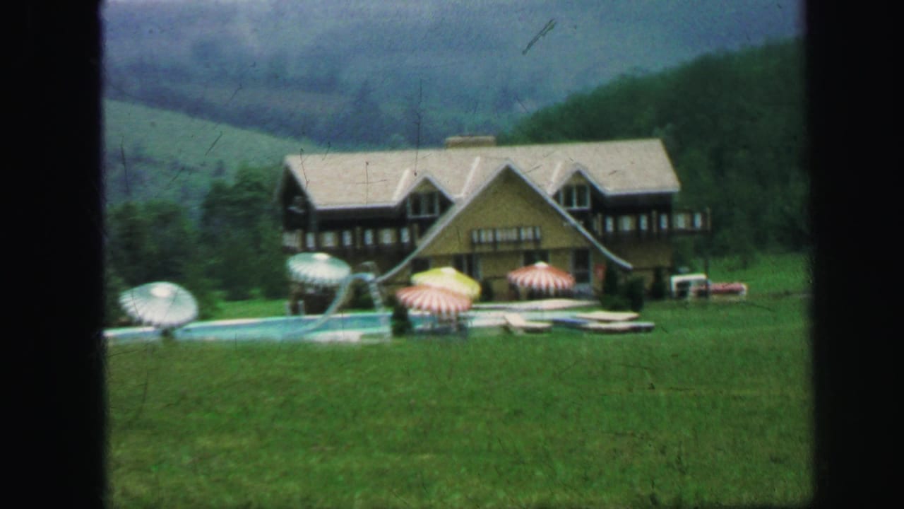 1971 8mm frame of a lodge-style building with pool, umbrellas, lawn, and wooded hills in the background.