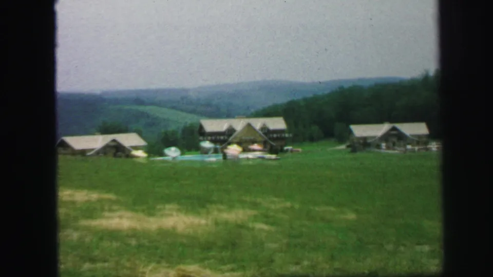 1971 8mm wide frame showing lodge buildings, cabins, poolside umbrellas, grass, and rolling hills.