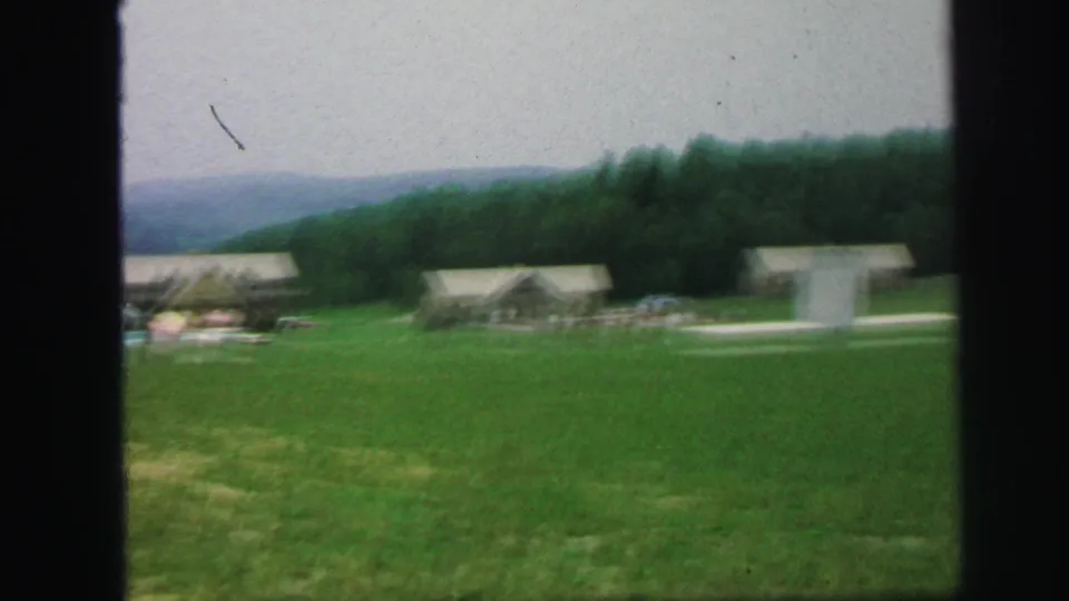 Soft 1971 8mm frame with cabins, open lawn, poolside area, and a pale vertical structure during the zoom or pan.