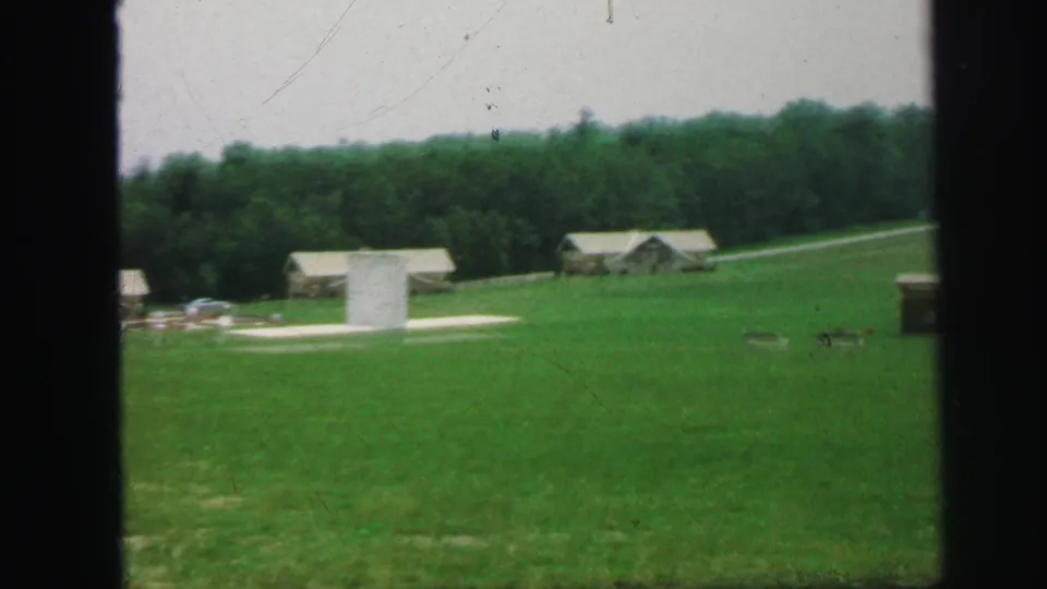 1971 8mm wide frame of resort-like grounds with cabins, grass, trees, a road, and a pale vertical structure.