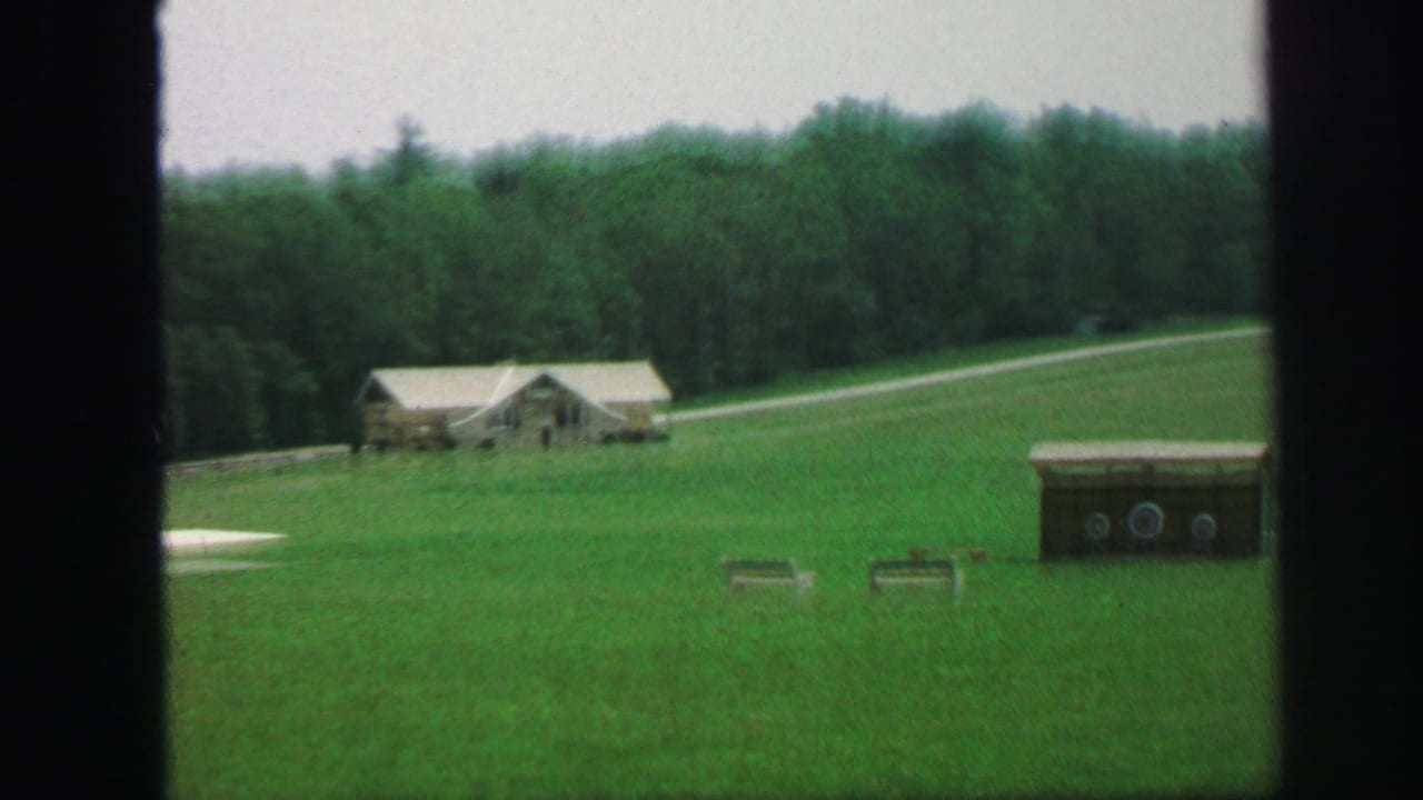 1971 Lynbrook, New York home-movie frame showing a green field, small house or outbuilding, treeline, gravel path, and target-like objects.