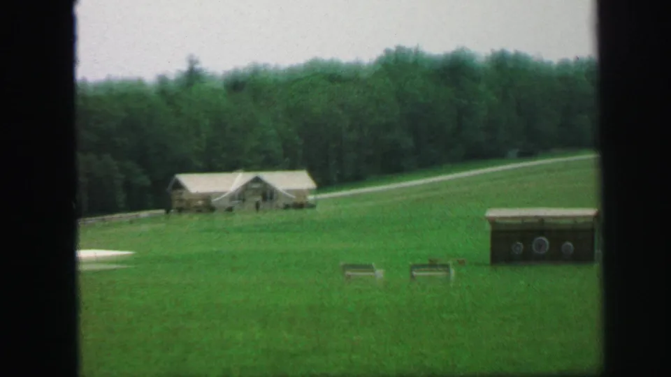 1971 Lynbrook, New York home-movie frame showing a green field, small house or outbuilding, treeline, gravel path, and target-like objects.