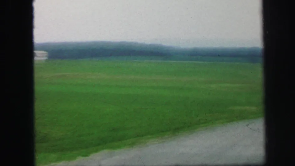 1971 8mm Lynbrook frame showing open green land, distant wooded horizon, muted overcast sky, and a gravel or paved edge at lower right.