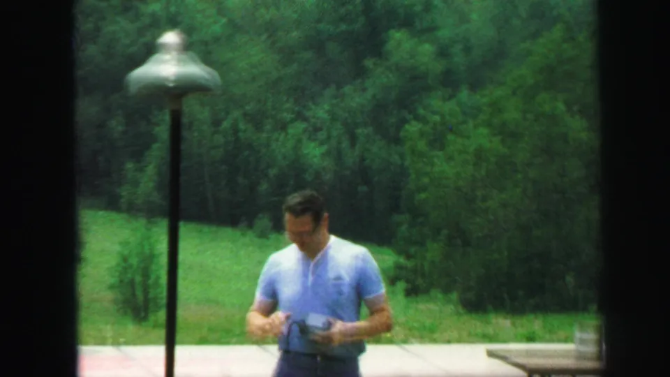 The man in a blue shirt stands near the outdoor table in Lynbrook, with a lamppost, lawn, and wooded backdrop visible.