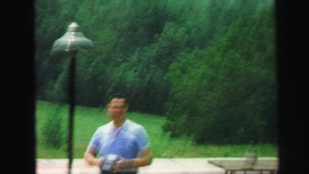 In 1971 Lynbrook home-movie footage, a man in a light blue shirt stands outdoors near a lamp post and green trees.
