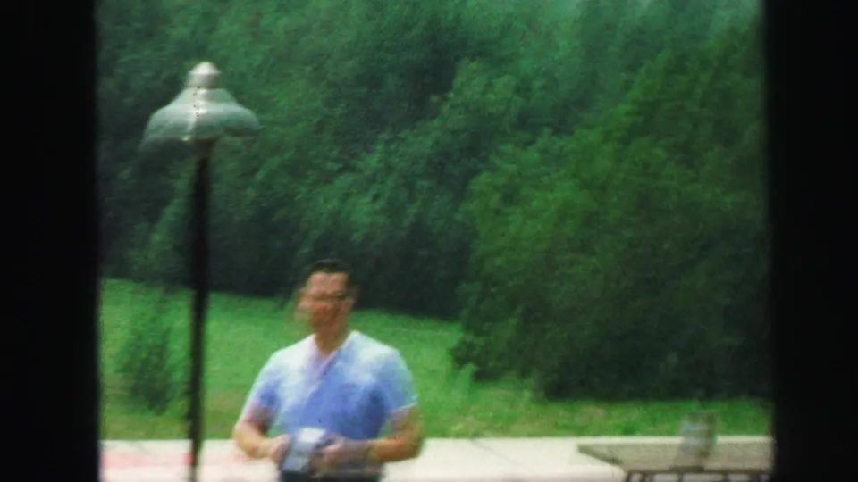 In 1971 Lynbrook home-movie footage, a man in a light blue shirt stands outdoors near a lamp post and green trees.