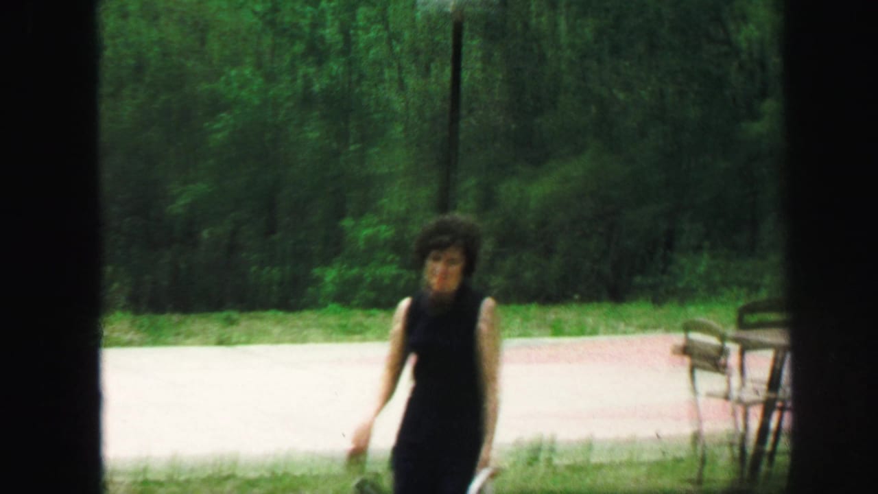 A woman in a dark sleeveless dress appears outdoors near a path and trees in Lynbrook, New York home-movie footage.