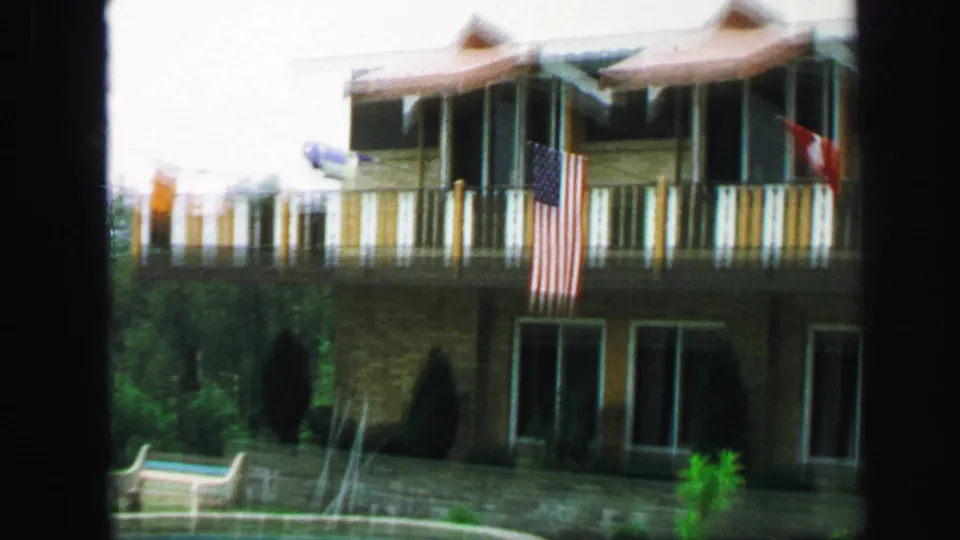A two-story Lynbrook residence appears with a balcony, American flag, striped trim, patio doors, shrubs, and chairs.
