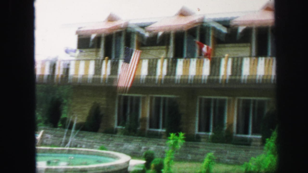 1971 Lynbrook home-movie frame showing a pool edge, yellow-brown building, balconies, striped railings, and American and Canadian flags.