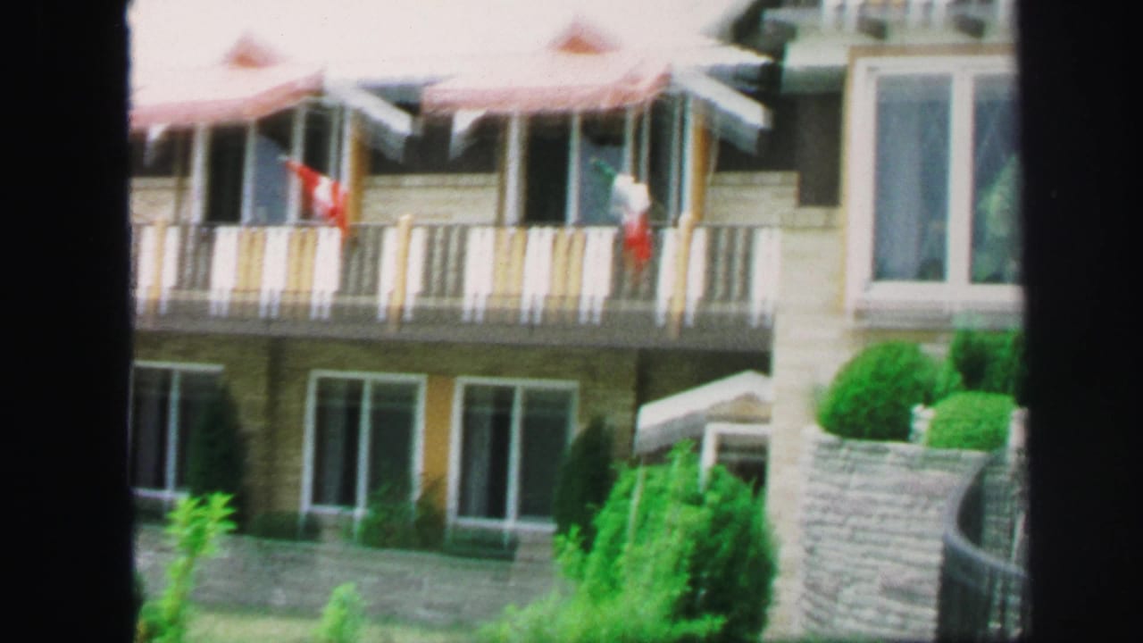 Balcony facade with flags, windows, shrubs, and a stone wall near the pool.