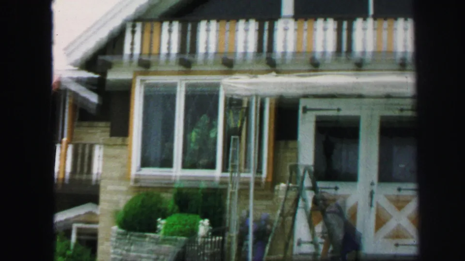 1971 Lynbrook home-movie frame showing a closer view of the yellow-brown building with balcony railings, bay window, entry doors, and trimmed shrubs.
