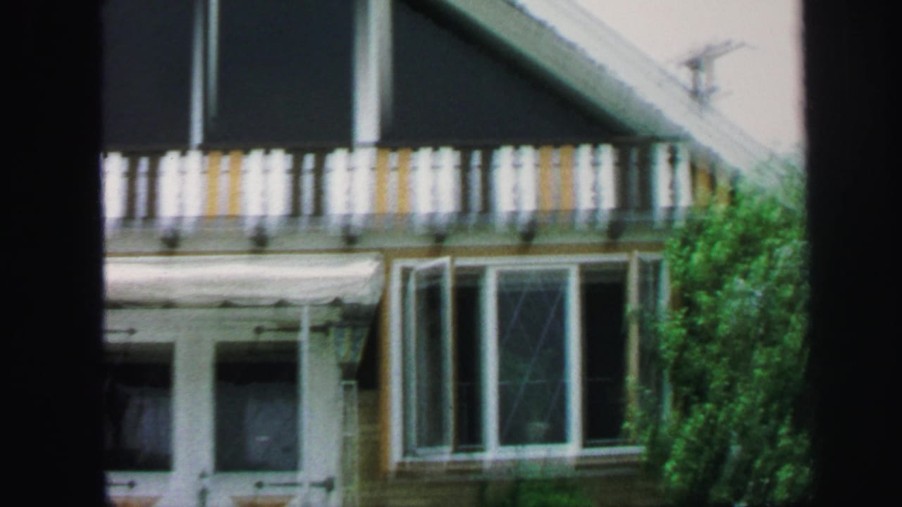 1971 Lynbrook home-movie frame showing a suburban house facade with a striped balcony, front windows, and dense green shrubs.