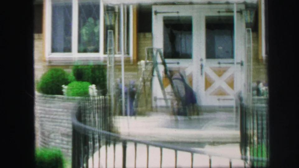 1971 Lynbrook home-movie view of the porch entrance, with white double doors, garden wall, fence, ladder, and blurred person nearby.