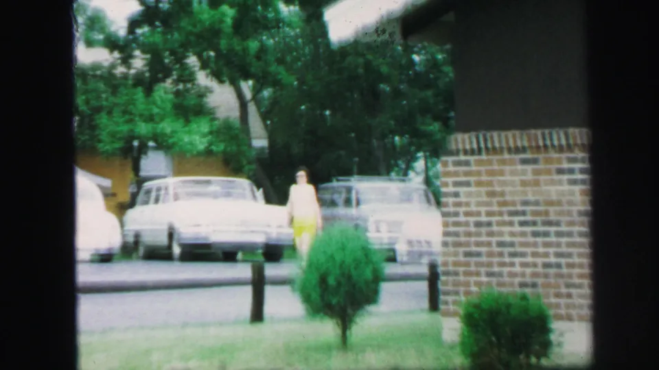 1971 Lynbrook street view with parked cars, a person in yellow walking past lawns, shrubs, trees, and a brick building edge.